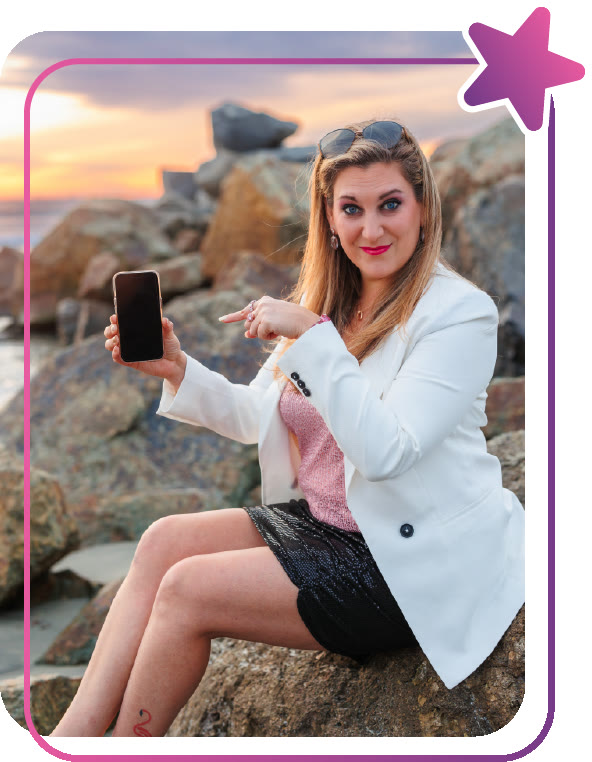 Amanda Robinson sitting on seaside rocks at sunset, pointing at her phone